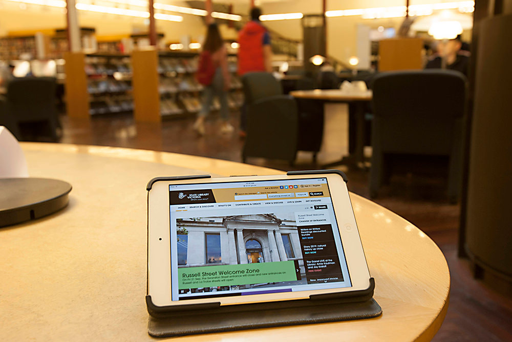 Tablet showing SLV home page shot on a round desk with bookshelves in background