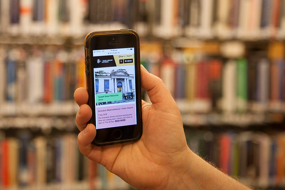 Hand holding an iphone showing SLV home page shot against shelves of books